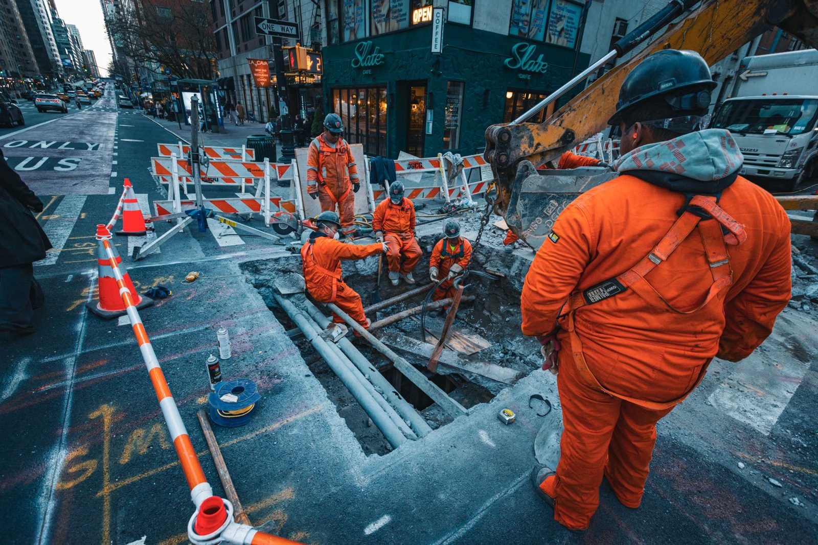 A crew in orange coveralls working a street trench behind barricades