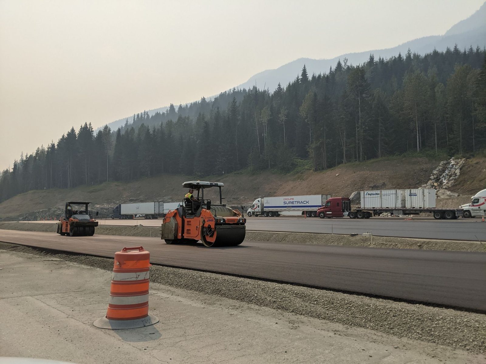 A paving crew rolling fresh asphalt on a mountain interstate lined with pine forest